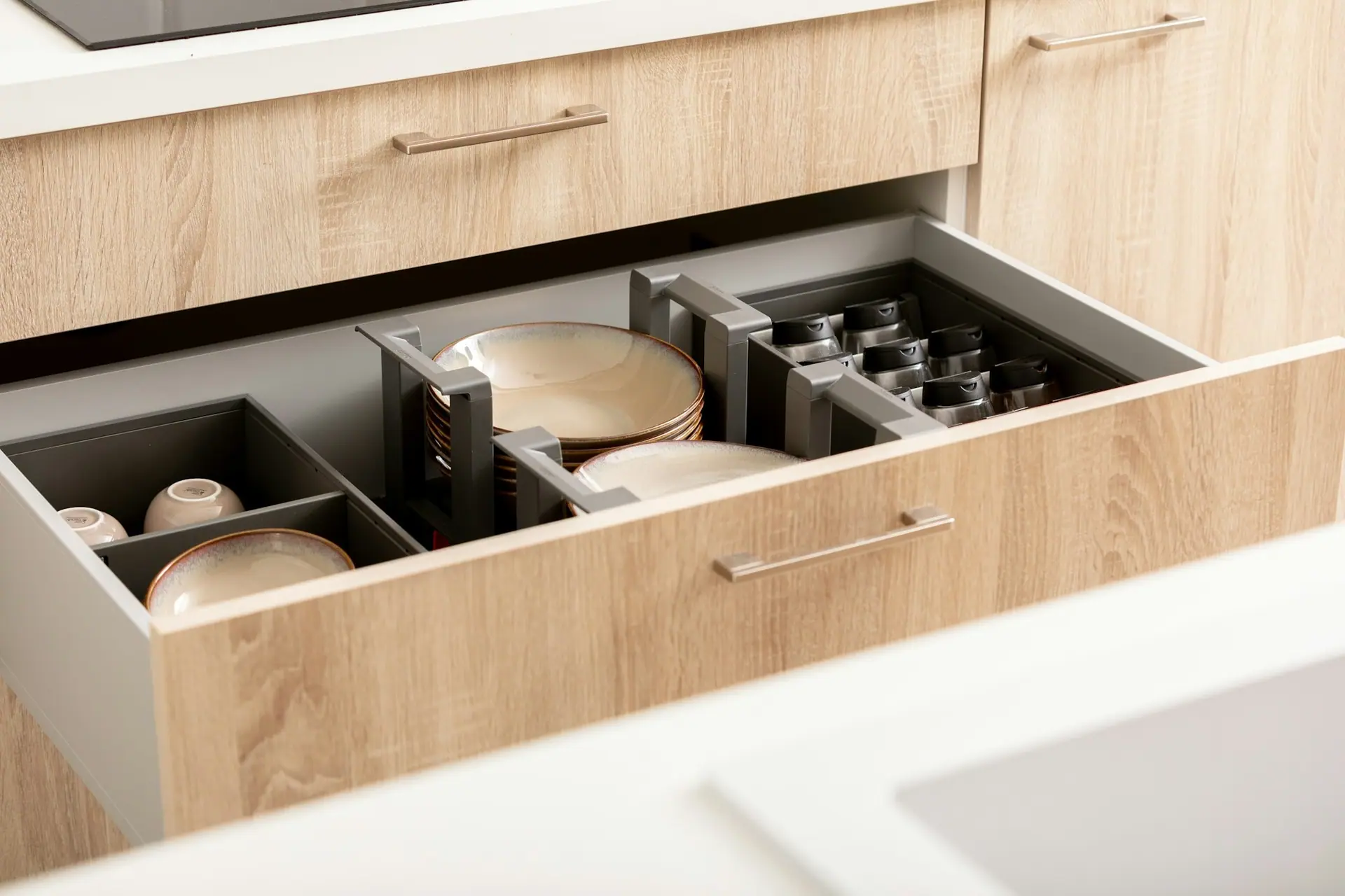 Open oak kitchen drawer with grey dividers holding plates and cups